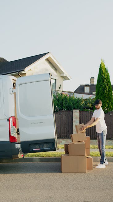 A man wearing a white t-shirt and jeans stands outside a residential property, preparing to load cardboard boxes into a white delivery van parked on the street. The boxes are stacked on the pavement near the open rear doors of the van, which reveal the interior used for transporting household items during a home relocation process. The van is positioned adjacent to a driveway with a small lawn and a wooden fence separating the property from neighboring houses. In the background, there is a white house with a pitched roof and some greenery, including a tall, conical-shaped shrub. The scene captures the logistical aspects of furniture transport and packing for a house move, with visible packing materials and the loading process in progress. Man with Van Islington provides services related to removals and moving logistics, as depicted in this unloading and loading scene.