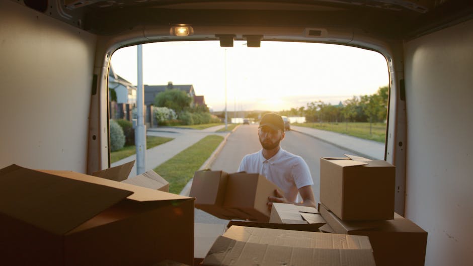 A man wearing a white polo shirt and baseball cap is seen through the open rear doors of a moving van during a home relocation process. Inside the van, there are several cardboard boxes of varying sizes, some partially open, arranged for transport or packing. The boxes are made of brown corrugated cardboard, with some wrapped in plastic or paper for added protection, and are positioned on the van floor, ready to be loaded or unloaded. The van's interior is illuminated by natural light from the outdoor environment, revealing a suburban street scene with a sidewalk, a lamppost, neatly trimmed bushes, and residential houses in the background. The setting appears to be during sunset, casting a warm glow over the scene. This image captures the loading or unloading phase of a furniture transport operation, illustrating a professional moving service like Man with Van Islington handling packing and logistics for an Upper Street move, as part of a comprehensive house removals process.