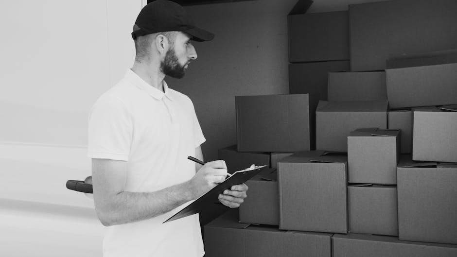 A man wearing a white t-shirt and jeans stands outside a residential property, preparing to load cardboard boxes into a white delivery van parked on the street. The boxes are stacked on the pavement near the open rear doors of the van, which reveal the interior used for transporting household items during a home relocation process. The van is positioned adjacent to a driveway with a small lawn and a wooden fence separating the property from neighboring houses. In the background, there is a white house with a pitched roof and some greenery, including a tall, conical-shaped shrub. The scene captures the logistical aspects of furniture transport and packing for a house move, with visible packing materials and the loading process in progress. Man with Van Islington provides services related to removals and moving logistics, as depicted in this unloading and loading scene.
