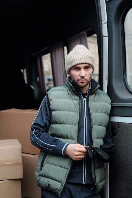 A man with a serious expression, wearing a light grey knitted beanie, a green and navy padded jacket, and dark gloves, stands inside a vehicle during a home relocation process. He is holding a black strap used for securing items during moving. Behind him, there are cardboard boxes stacked, indicating packing for a move. The vehicle's interior is dark, with a window partially visible, showing a glimpse of the exterior environment. This scene illustrates a professional removals service, possibly provided by Man with Van Islington, focused on furniture and household item transport as part of a moving and packing process.