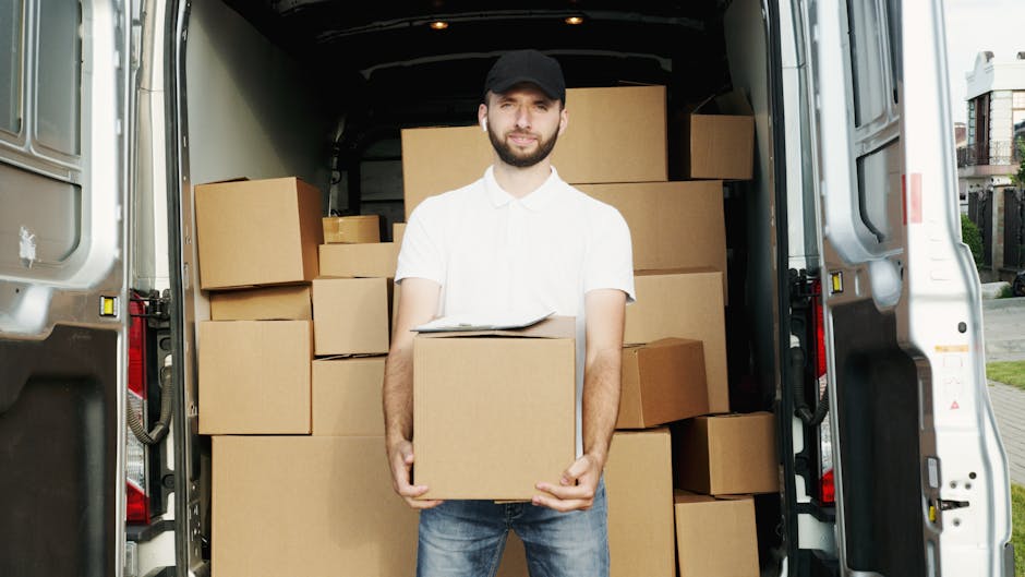 A man with a beard, wearing a black cap, white polo shirt, and blue jeans, stands in front of the open rear doors of a moving van filled with neatly stacked, medium-sized cardboard boxes wrapped in plastic and secured with packing materials. He holds a single box in his hands, with additional boxes visible inside the van, some stacked against the walls. The van is parked outside on a paved area with a residential street and houses in the background. The lighting indicates natural daylight, and the interior of the van shows a loading process typical of home relocation or furniture transport, with packaging and moving equipment such as straps or blankets possibly present. This scene demonstrates professional removals carried out by Man with Van Islington, reflecting the packing and loading stage of house removals services.