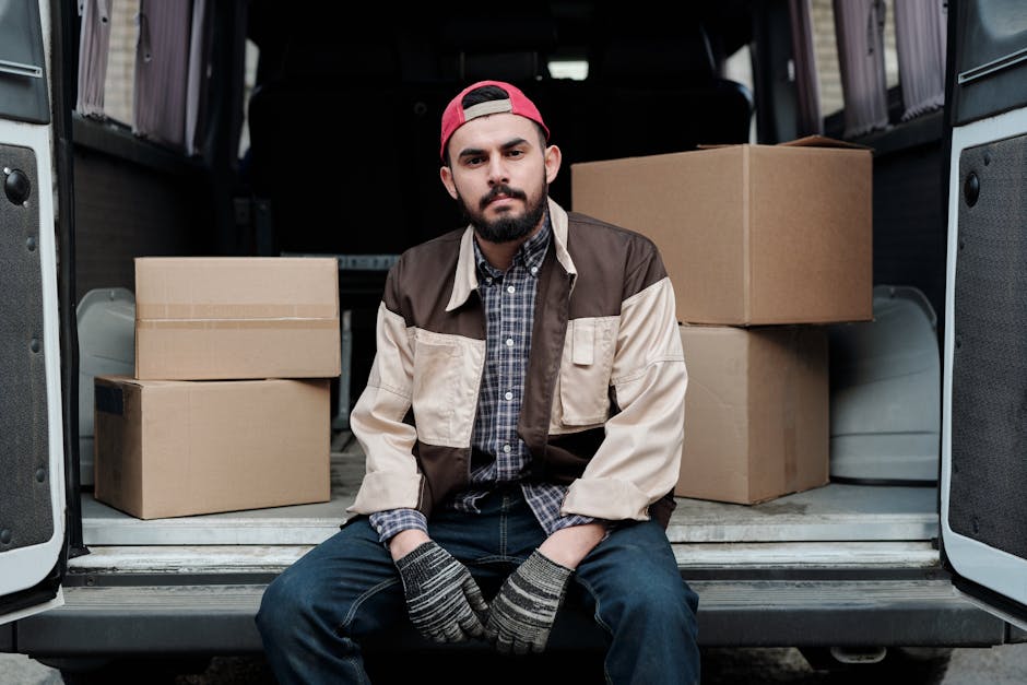 A man with a serious expression, wearing a light grey knitted beanie, a green and navy padded jacket, and dark gloves, stands inside a vehicle during a home relocation process. He is holding a black strap used for securing items during moving. Behind him, there are cardboard boxes stacked, indicating packing for a move. The vehicle's interior is dark, with a window partially visible, showing a glimpse of the exterior environment. This scene illustrates a professional removals service, possibly provided by Man with Van Islington, focused on furniture and household item transport as part of a moving and packing process.