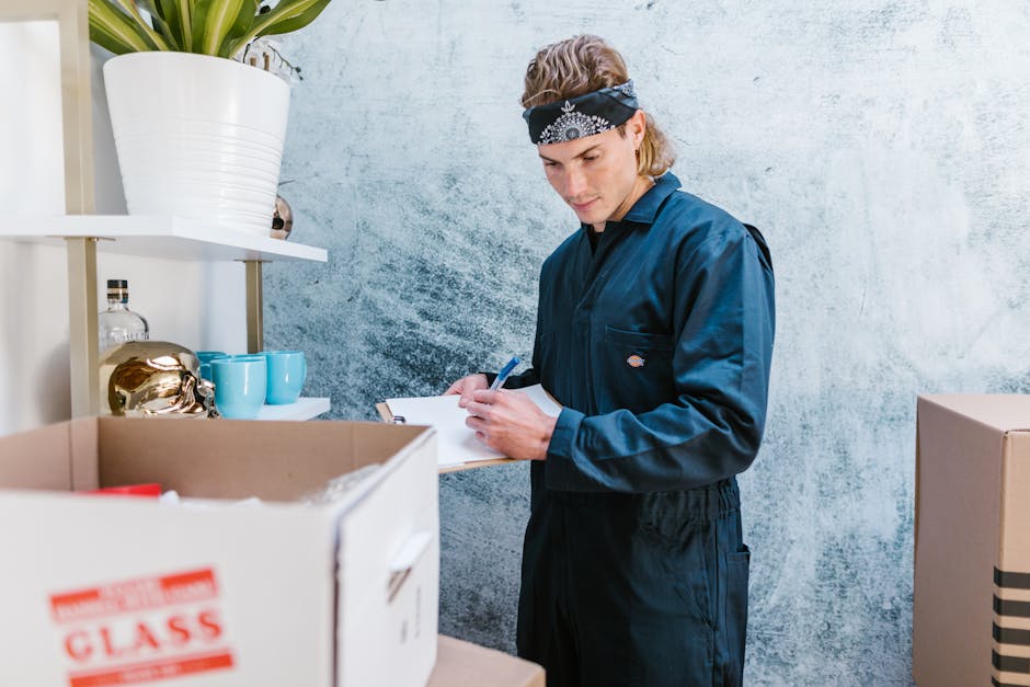 A young male mover, dressed in a dark blue uniform and wearing a black bandana, is inside a room with a textured light grey wall. He is holding a pen and a clipboard, documenting items for a house removal. In front of him, there are open cardboard boxes filled with household items, including some cardboard packing materials. To the left, a white shelving unit supports decorative objects such as a white ceramic pot with a plant, a gold decorative piece, and several blue cups. The setting appears to be a preparation area for a home relocation, with furniture and packing materials arranged around. The image illustrates the packing and moving process, with the professional from Man with Van Islington meticulously recording inventory as part of the house removals service for the Canonbury area, supporting efficient furniture transport and logistics for the upcoming move.