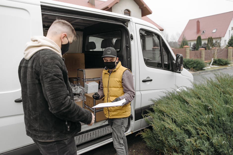 Two men wearing black face masks are engaged in a home relocation process involving furniture and packing supplies outside a white van parked in a residential area with houses and greenery. The man on the left, dressed in a black jacket and grey trousers, is holding a clipboard or document while the man on the right, wearing a yellow puffer vest over a grey hoodie and a black cap, appears to be reviewing the paperwork. Inside the van, visible cardboard boxes and plastic-wrapped items are being prepared for loading or unloading. The scene is lit with natural daylight, and the van is positioned near a driveway or street, with shrubs and a row of houses with sloped roofs in the background. The image reflects the careful packing and moving logistics typical of house removals, with bedding, packaging materials, and furniture likely being part of the transportation process managed by Man with Van Islington, supporting scheduled home relocations around Barnsbury streets.