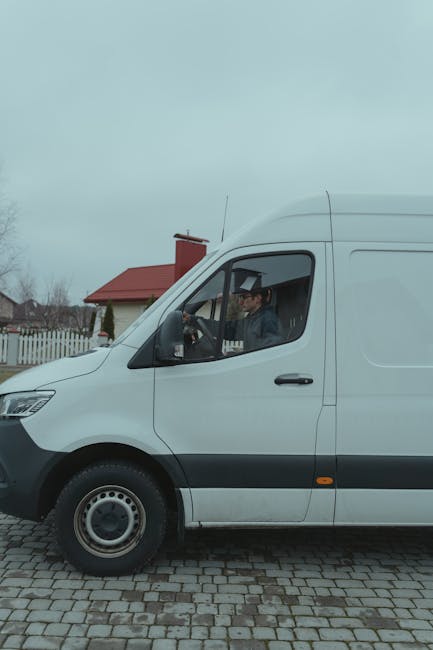 A white panel van with a sliding side door is parked on a cobblestone driveway in front of a residential house with a red roof and white picket fence. Inside the van, a person wearing a dark jacket and cap is visible through the open driver's side window, appearing to be preparing for a home relocation or furniture transport. The scene is set outdoors during daylight hours with overcast skies, and the environment suggests the beginning or ongoing phase of moving household items, consistent with house removals services. Man with Van Islington's service focus on moving logistics is supported by the presence of the vehicle and the loading area, emphasizing the transportation phase of a domestic move.