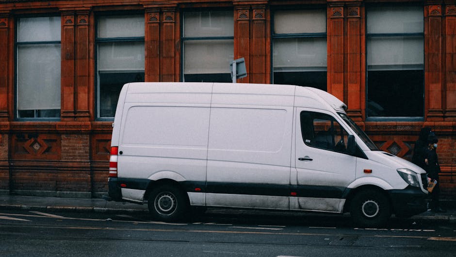 A white panel van with a sliding side door is parked on a cobblestone driveway in front of a residential house with a red roof and white picket fence. Inside the van, a person wearing a dark jacket and cap is visible through the open driver's side window, appearing to be preparing for a home relocation or furniture transport. The scene is set outdoors during daylight hours with overcast skies, and the environment suggests the beginning or ongoing phase of moving household items, consistent with house removals services. Man with Van Islington's service focus on moving logistics is supported by the presence of the vehicle and the loading area, emphasizing the transportation phase of a domestic move.