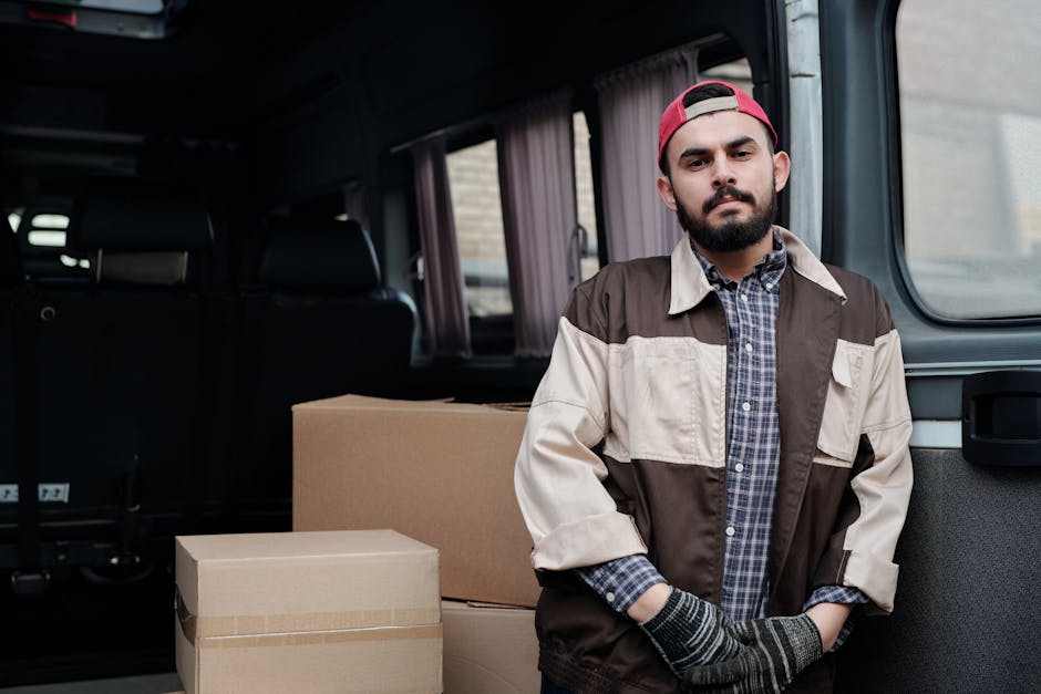 A man dressed in a checked shirt, beige and brown jacket, and a red cap stands inside a moving van, positioned near the open rear door. In front of him are several cardboard boxes of varying sizes, so
