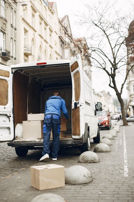 A man wearing a blue jacket and dark trousers is standing inside the open rear cargo space of a white moving van, which is parked on a city street with residential buildings visible in the background. The van's interior shows wooden paneling and is partially filled with white cardboard boxes and packaging materials. Outside the van, a cardboard box rests on the paved pavement nearby, with large rounded concrete bollards separating the parking area from the street. The scene suggests the process of home relocation, with the man loading or unloading items as part of furniture transport or packing and moving activities. The street is lined with leafless trees and other parked vehicles, and the overall environment appears calm and organized, in line with regulations for removals and loading areas, as handled by [COMPANY_NAME].