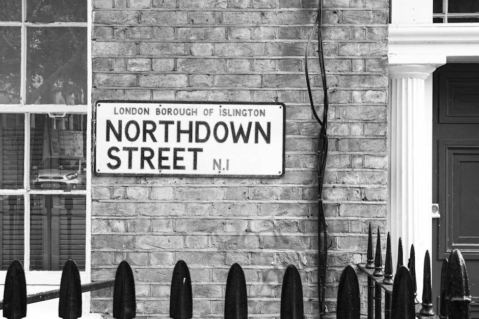 A black and white photograph of a residential street scene in Islington, showing a brick building with a street sign that reads 'Northdown Street, London Borough of Islington, N1'. The sign is mounted on the brick wall to the left of a large window with white framing, which reveals reflections of parked cars and the street outside. To the right of the sign, electrical cables are attached to the wall, hanging loosely. A partially visible door with decorative columns is adjacent to the cables. In the foreground, a black metal fence with pointed finials runs along the pavement, enclosing the front yard or entrance area of the property. The area suggests an urban setting where a house removal or moving process, involving packing, furniture transport, or loading activities, could take place, with nearby parking restrictions or permissions relevant to such activities. Man with Van Islington often manages the logistics of home relocation, including loading and unloading of furniture and packing materials like cardboard boxes, plastic wrap, and blankets.