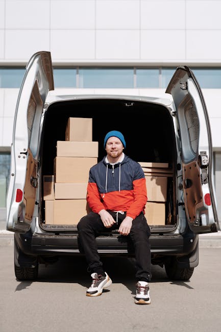 A man wearing a blue jacket and dark trousers is standing inside the open rear cargo space of a white moving van, which is parked on a city street with residential buildings visible in the background. The van's interior shows wooden paneling and is partially filled with white cardboard boxes and packaging materials. Outside the van, a cardboard box rests on the paved pavement nearby, with large rounded concrete bollards separating the parking area from the street. The scene suggests the process of home relocation, with the man loading or unloading items as part of furniture transport or packing and moving activities. The street is lined with leafless trees and other parked vehicles, and the overall environment appears calm and organized, in line with regulations for removals and loading areas, as handled by [COMPANY_NAME].