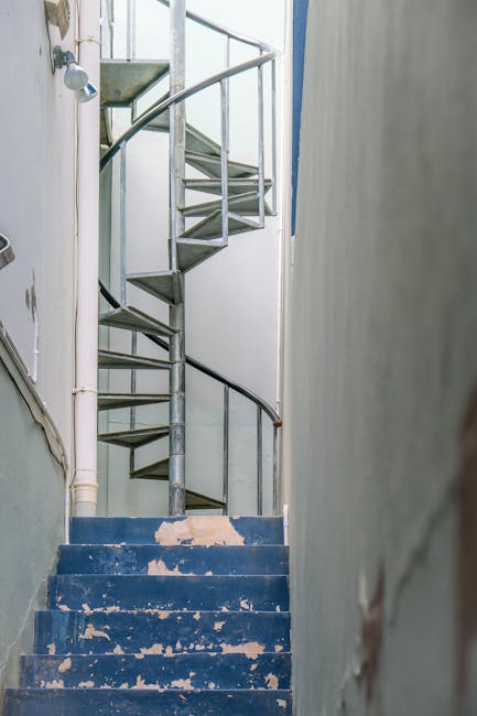An interior view of a narrow, worn staircase with blue-painted steps that are chipped and peeling, leading up to a small landing with a metal spiral staircase ascending further. The metal staircase has a black handrail and silver steps, positioned against a white wall, which shows signs of age and minor damage. To the left, there are exposed white pipes running vertically along the wall. The setting appears to be inside a residential building, prepared for a home relocation or furniture transport, with natural light illuminating the space. The image emphasizes challenging access conditions typical of tight staircases in urban flats requiring careful handling during moving and packing processes, highlighting the need for professional removals services such as those offered by Man with Van Islington.