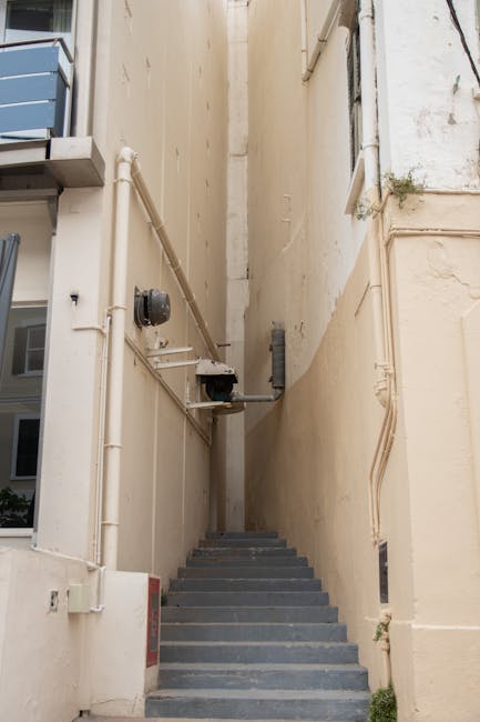 A narrow outdoor staircase with dark grey steps leads between two high beige building walls, which are fitted with various utility pipes, including white PVC pipes, a black meter, and electrical conduits. The walls are plain with some minor discoloration and have small plants growing from cracks. To the left, part of a modern building with blue window frames and balconies is visible. The area appears to be an alleyway or back access route, demonstrating the challenging space for home removal or furniture transport. The scene underscores the logistical considerations involved in moving items through tight access points, typical of flat relocations in urban environments, as managed by companies like Man with Van Islington.