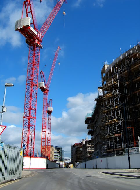 An image showing a construction site with three large red tower cranes lifting construction materials beneath a partly cloudy blue sky. To the right, a multi-storey building is under construction, sur