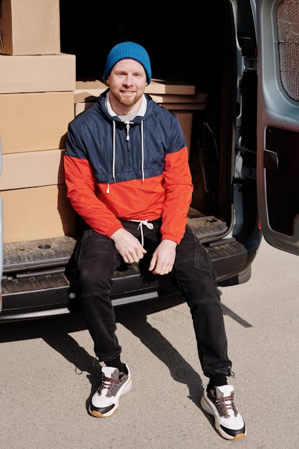 A man with light skin, wearing a blue beanie hat and a navy and red jacket, is seated on the rear bumper of a black van that has its side door open, revealing a loading area filled with stacked cardboard boxes and wrapped furniture. The boxes vary in size and are secured with packing tape, indicating a home relocation process. The man is positioned outdoors on a paved surface, possibly a driveway or street, with sunlight illuminating the scene. Inside the van, visible behind the man, are wooden furniture pieces and plastic-wrapped items being prepared for transport. The loading process appears organized, with the man ready to assist with the furniture transport or packing and moving tasks as part of a professional removal service, consistent with services offered by Man with Van Islington, specializing in removals and moving logistics.