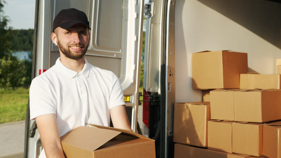 A man with light skin, wearing a blue beanie hat and a navy and red jacket, is seated on the rear bumper of a black van that has its side door open, revealing a loading area filled with stacked cardboard boxes and wrapped furniture. The boxes vary in size and are secured with packing tape, indicating a home relocation process. The man is positioned outdoors on a paved surface, possibly a driveway or street, with sunlight illuminating the scene. Inside the van, visible behind the man, are wooden furniture pieces and plastic-wrapped items being prepared for transport. The loading process appears organized, with the man ready to assist with the furniture transport or packing and moving tasks as part of a professional removal service, consistent with services offered by Man with Van Islington, specializing in removals and moving logistics.
